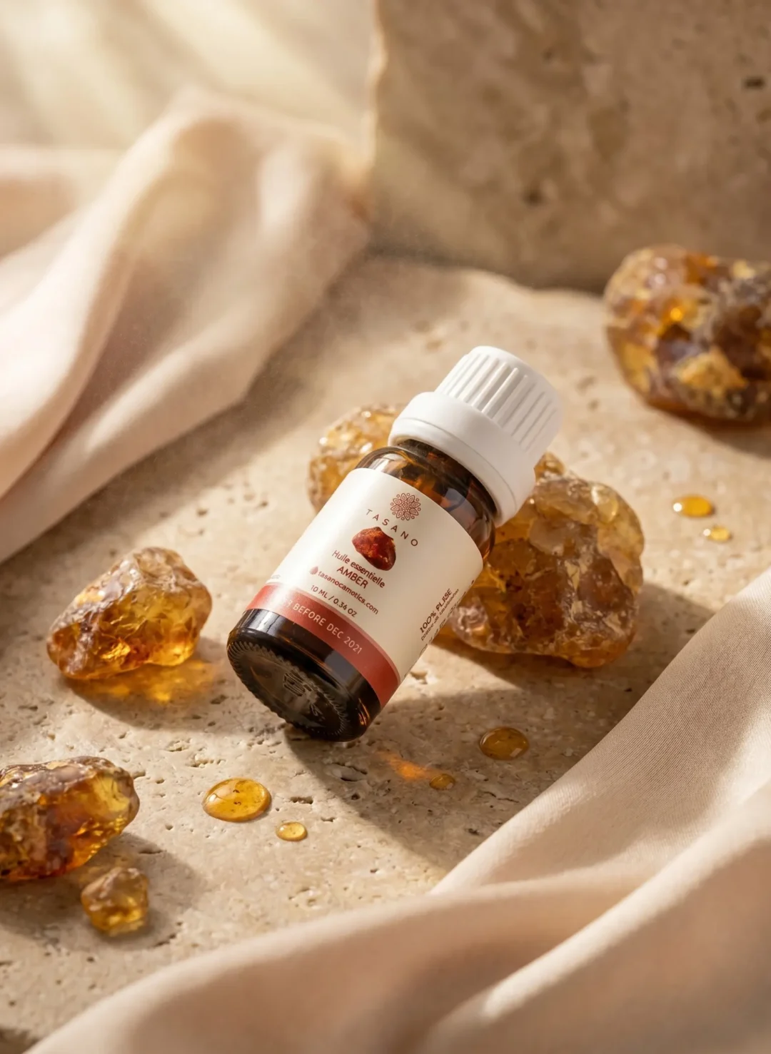 TASANO Amber Essential Oil styled with raw amber resin, warm oil drops, silk fabric, and natural sunlight on beige stone.