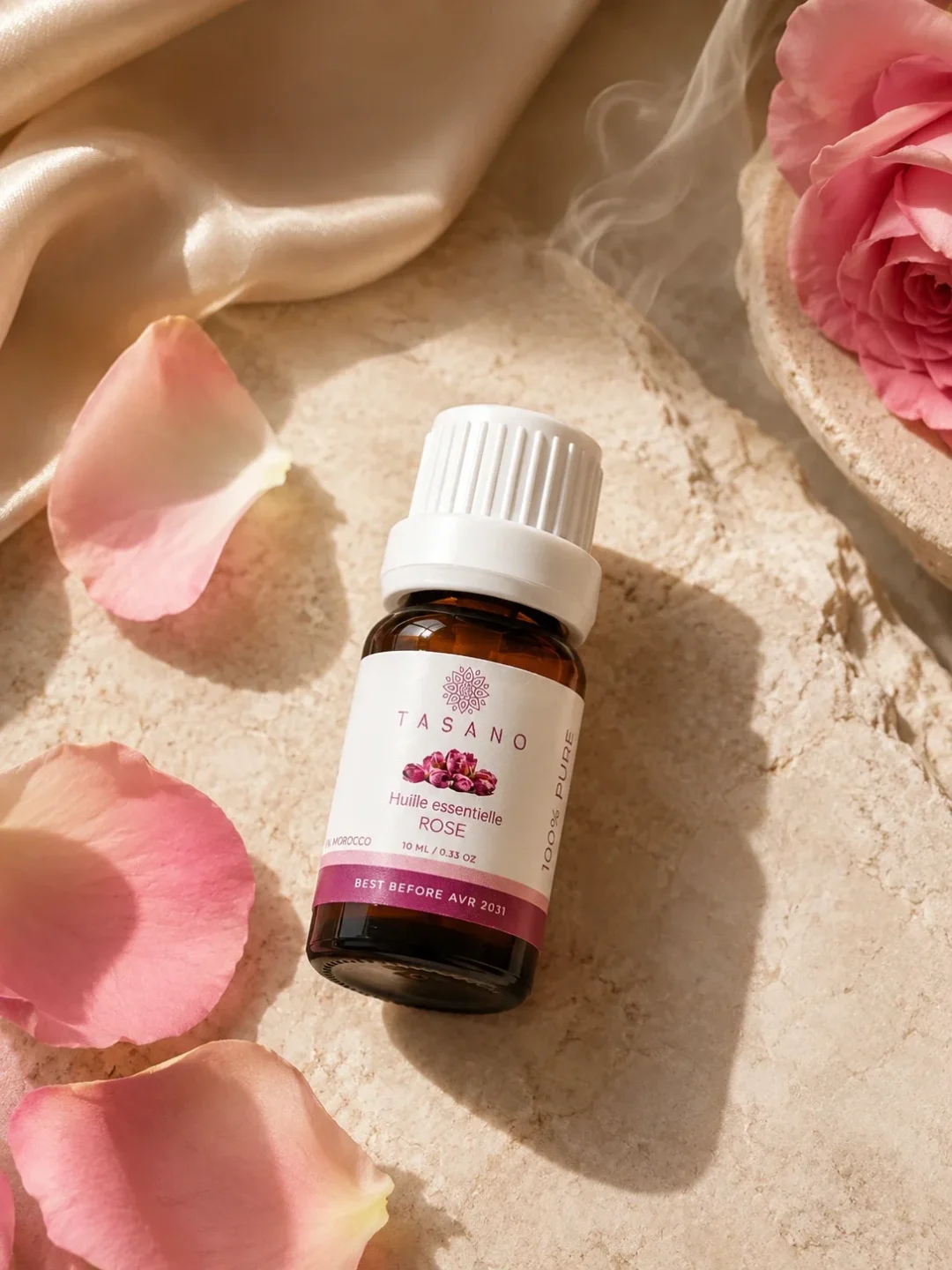 TASANO Rose Essential Oil styled with fresh rose petals, silk fabric, subtle vapour, and natural sunlight on beige stone.