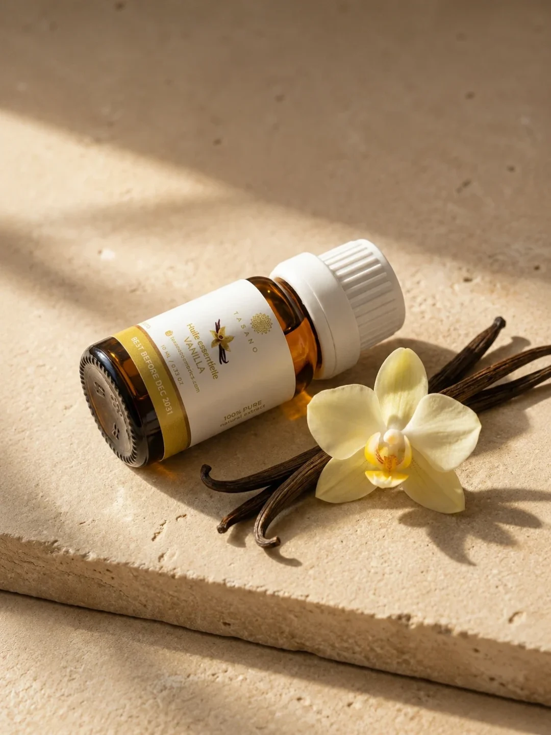TASANO Vanilla Essential Oil styled with vanilla beans, vanilla flower, golden oil drops, and sunlit beige stone.